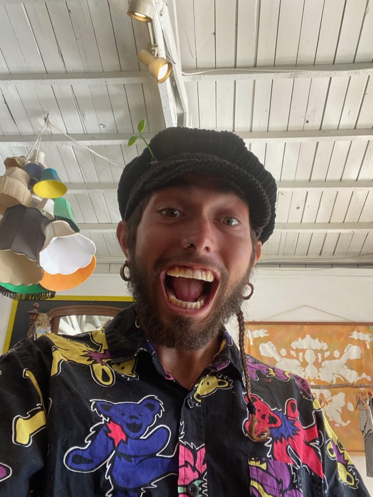 A person with a beard and a nose ring is smiling widely at the camera, wearing a colorful shirt and a knit cap, indoors with white ceiling.